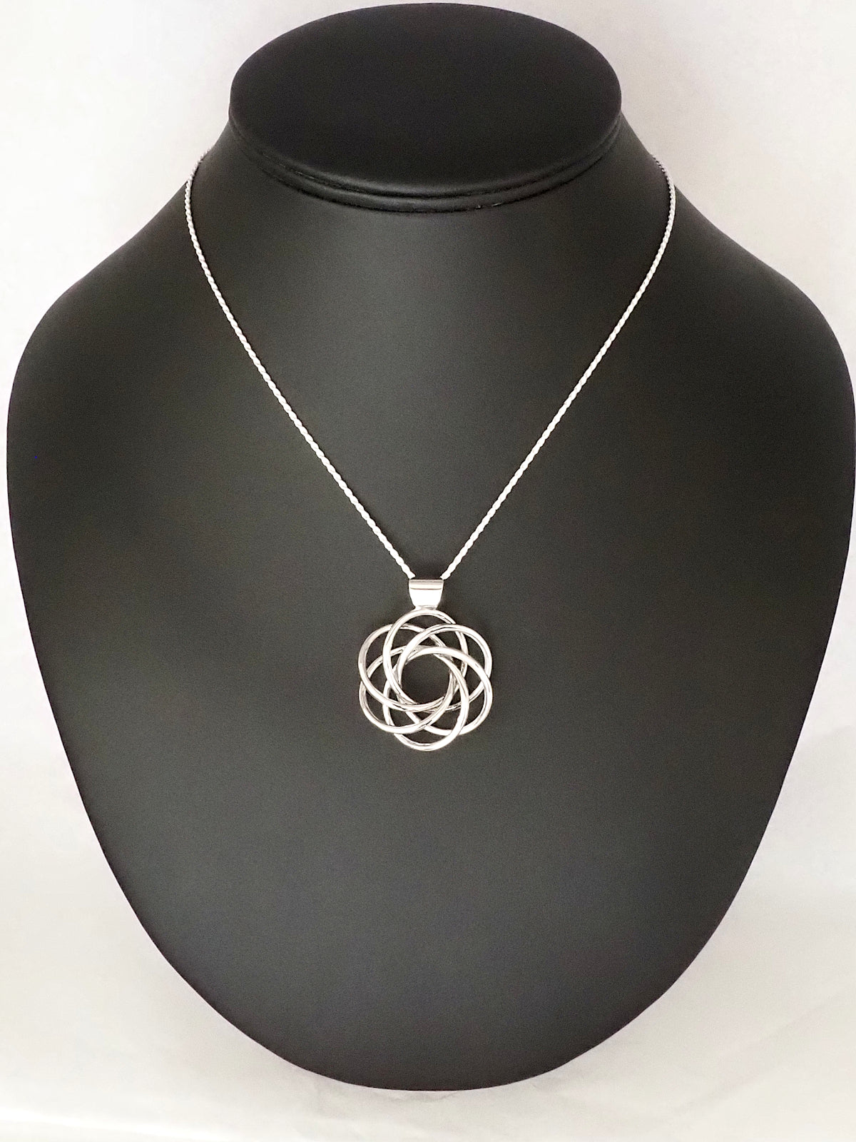 A  rhodium-plated brass pendant in an abstract shape resembling a flower with six petals, with a silver French rope chain, on a necklace display.