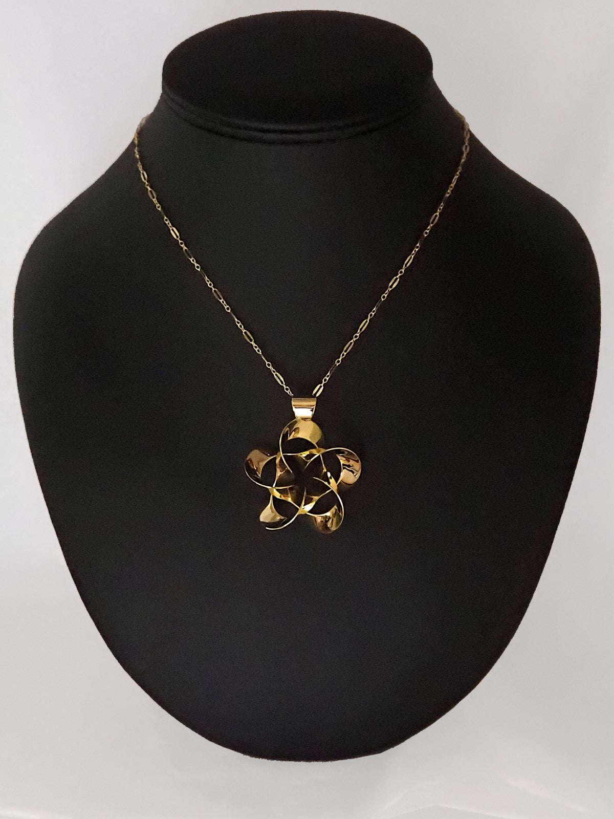 A shiny, gold-plated brass pendant in a flower-like shape with five petals, on a black display stand.