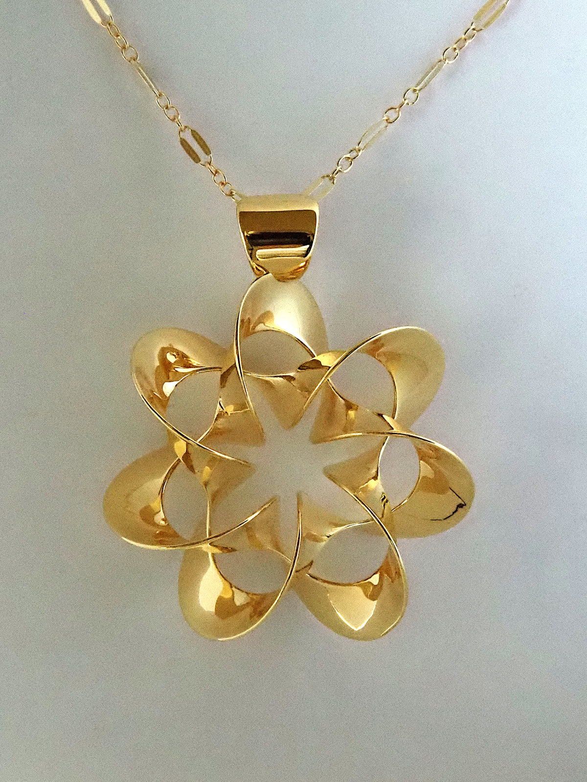 A shiny, gold-plated brass pendant in a flower-like shape with seven petals, on a white background.