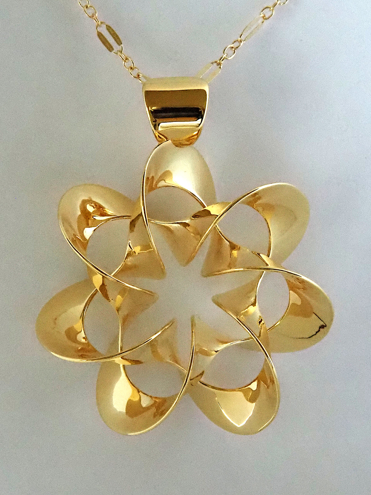 A shiny, gold-plated brass pendant in a flower-like shape with seven petals, on a white background.