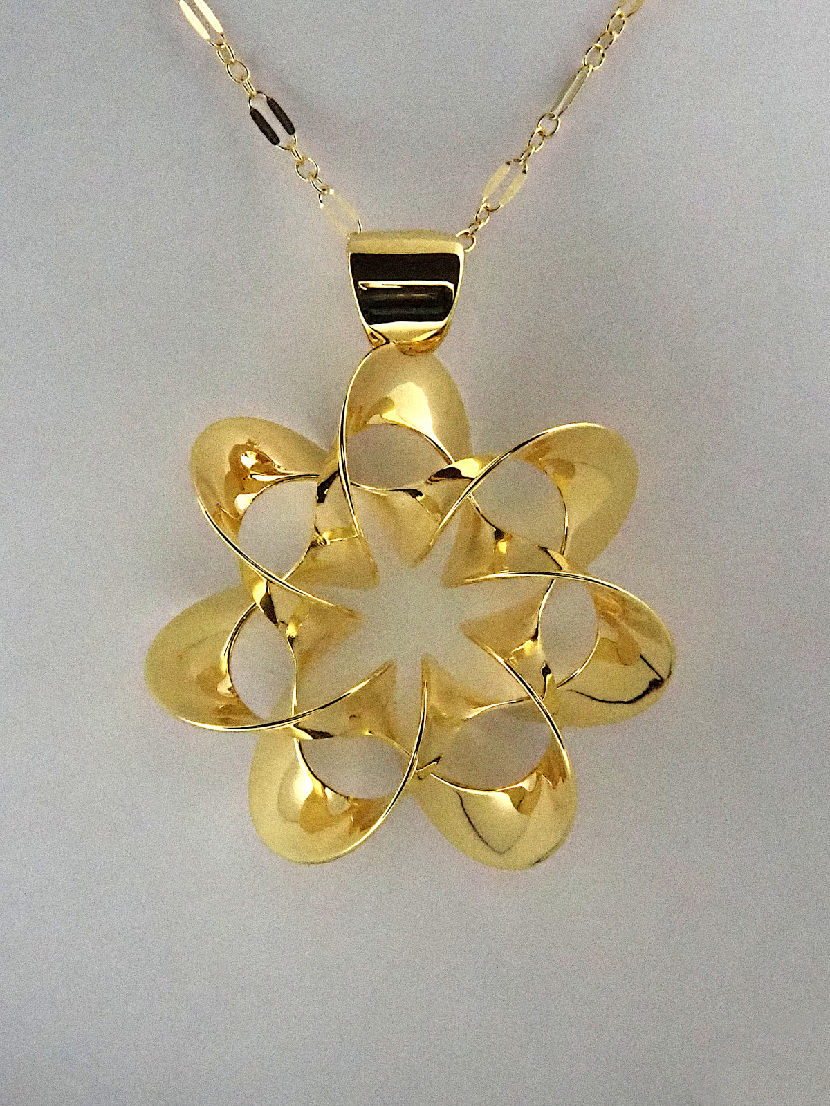 A shiny, gold-plated brass pendant in a flower-like shape with seven petals, on a white background.