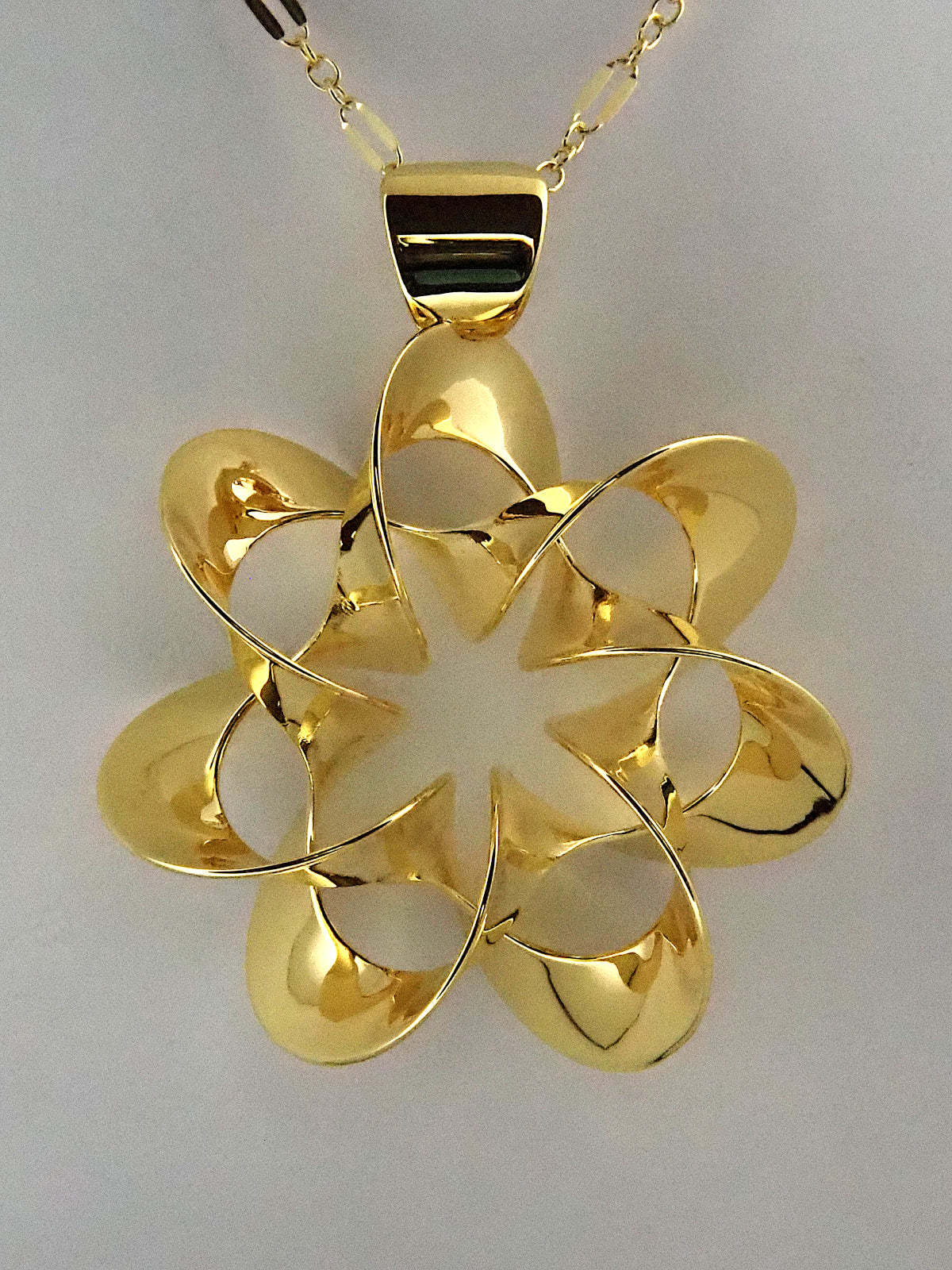 A shiny, gold-plated brass pendant in a flower-like shape with seven petals, on a white background.