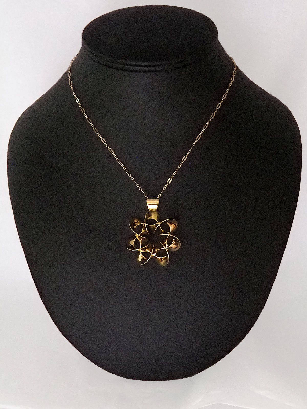 A shiny, gold-plated brass pendant in a flower-like shape with seven petals, on a black display stand.