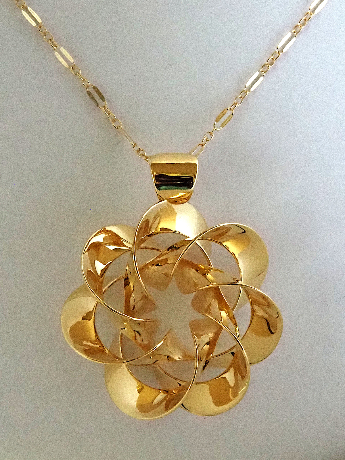 A shiny, gold-plated brass pendant in a flower-like shape with seven petals, on a white background.