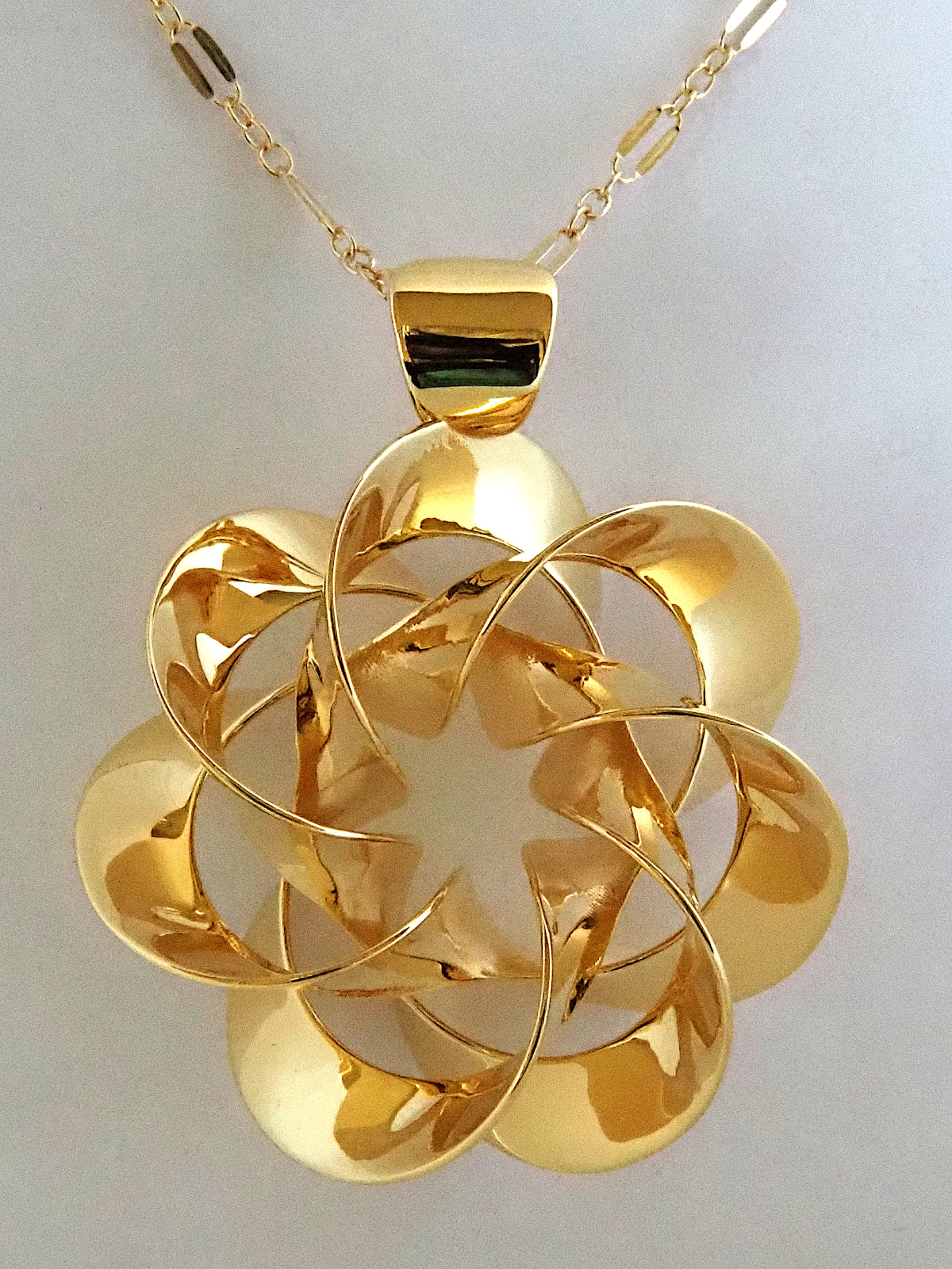 A shiny, gold-plated brass pendant in a flower-like shape with seven petals, on a white background.