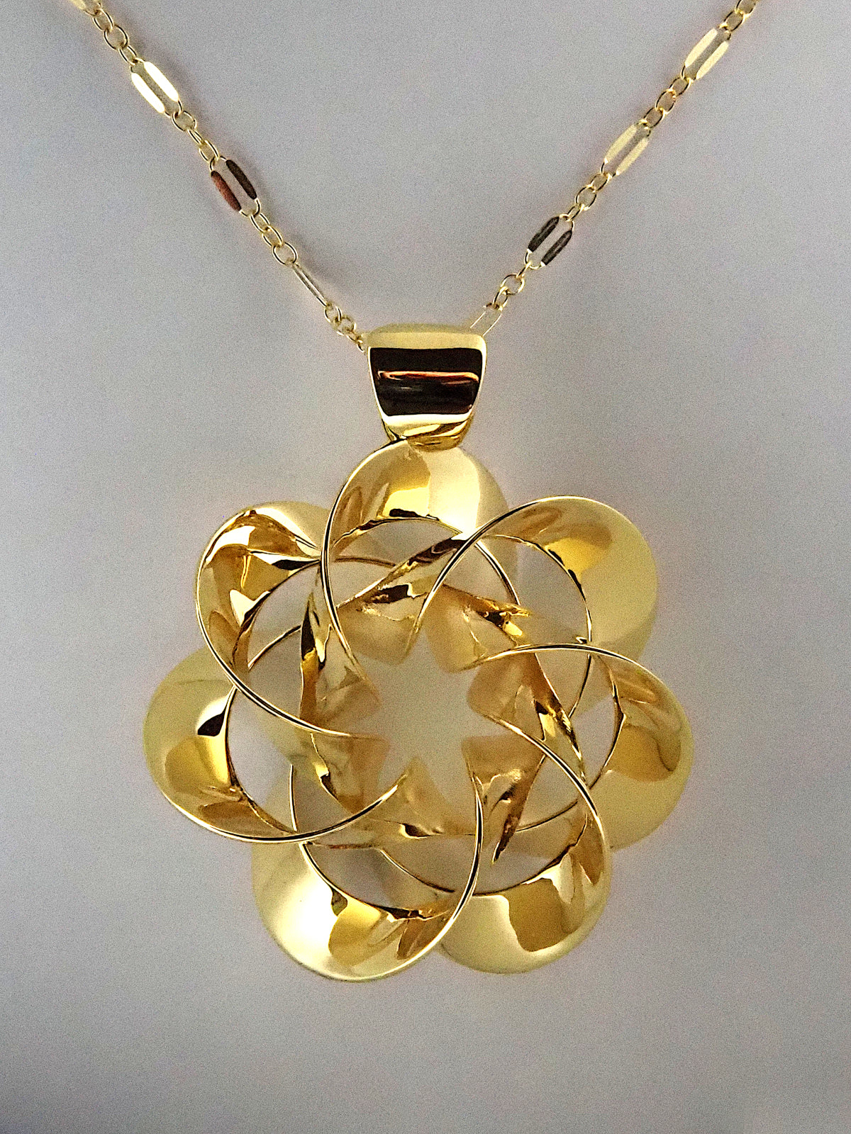 A shiny, gold-plated brass pendant in a flower-like shape with seven petals, on a white background.