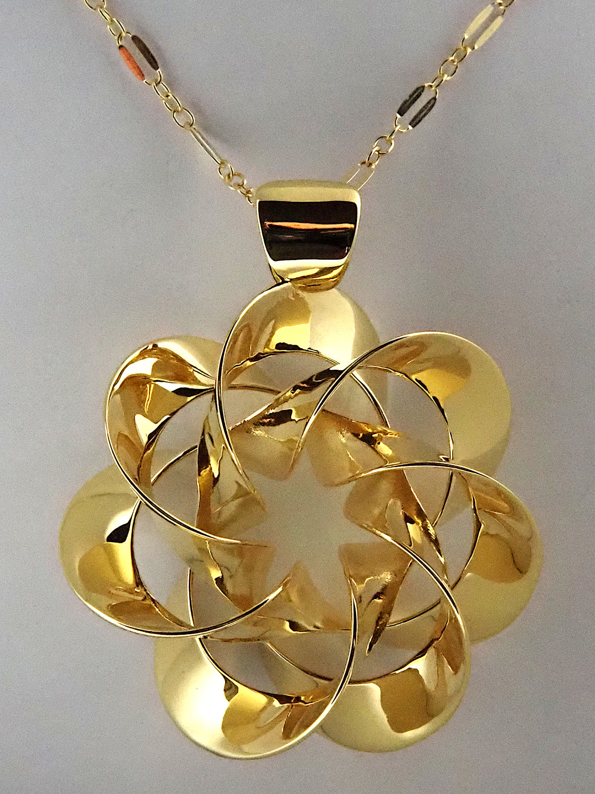 A shiny, gold-plated brass pendant in a flower-like shape with seven petals, on a white background.