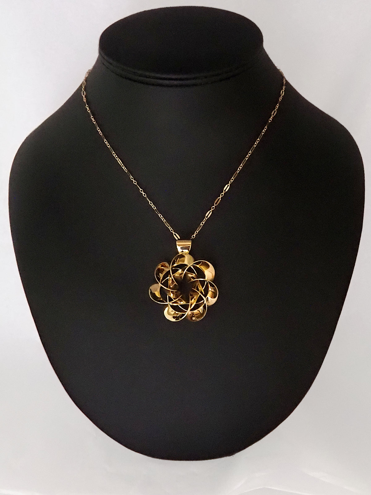 A shiny, gold-plated brass pendant in a flower-like shape with seven petals, on a black display stand.