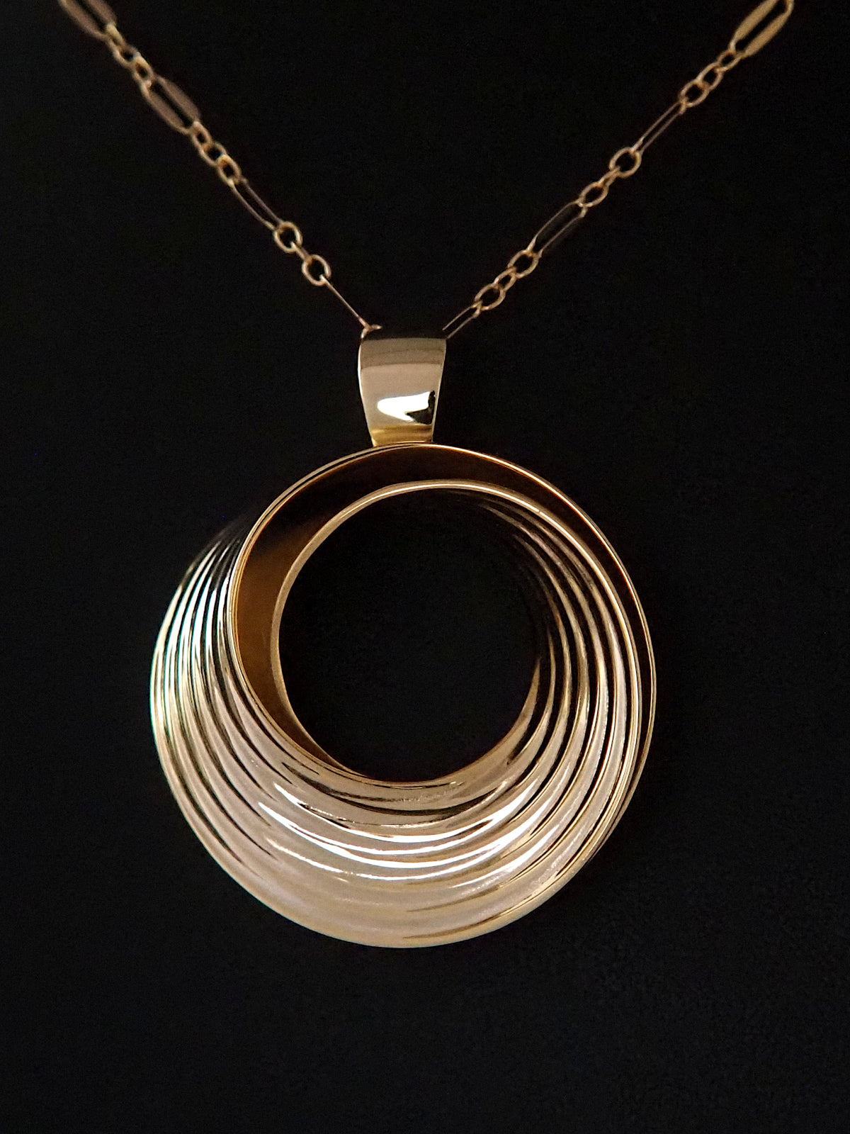 Photograph of a round, gold-plated silver pendant in the shape of a Moebius strip split into two layers, and ridged on the outside, on a gold long-and-short loop chain.