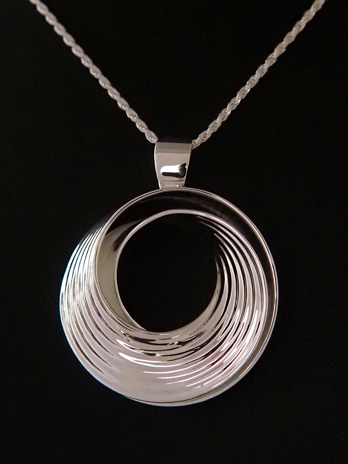 Photograph of a round, polished sterling silver pendant in the shape of a Moebius strip split into two layers, and ridged on the outside, on a sterling silver rope chain.