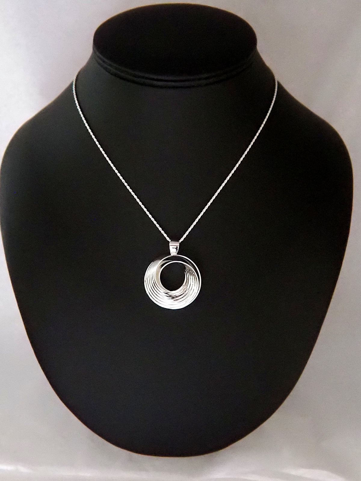 Photograph of a round, polished sterling silver pendant in the shape of a Moebius strip split into two layers, and ridged on the outside, on a sterling silver rope chain.