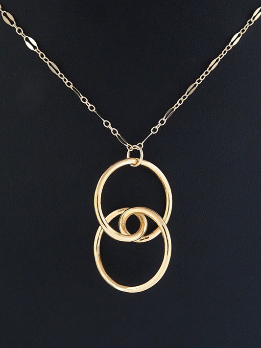 A gold-plated brass trefoil knot pendant on a black background in the general shape of two linked circular rings whose intersection is a stylized eye and having two-fold rotational symmetry about its horizontal and vertical axes, and about its center.