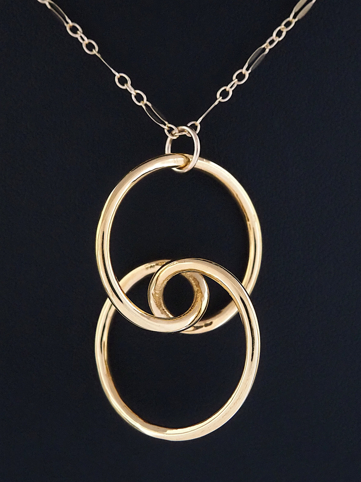 Close-up of a gold-plated brass trefoil knot pendant on a black background in the general shape of two linked circular rings whose intersection is a stylized eye and having two-fold rotational symmetry about its horizontal and vertical axes, and about its center.