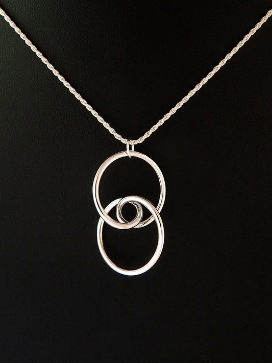 A rhodium-plated brass trefoil knot pendant on a black background in the general shape of two linked circular rings whose intersection is a stylized eye and having two-fold rotational symmetry about its horizontal and vertical axes, and about its center.