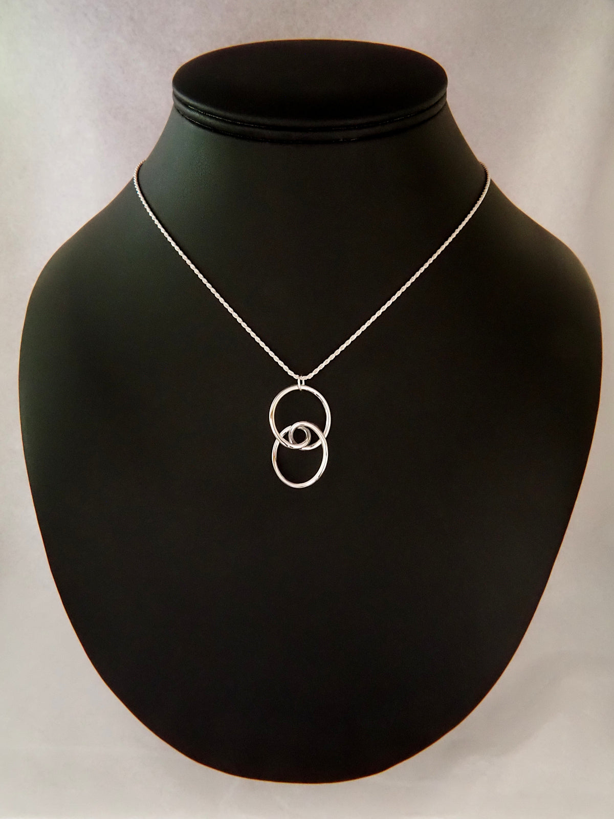 A rhodium-plated brass trefoil knot pendant on a black background in the general shape of two linked circular rings whose intersection is a stylized eye and having two-fold rotational symmetry about its horizontal and vertical axes, and about its center.